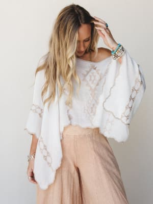 A breezy white poncho top with tan geometric embroidery and scalloped edges is paired with soft blush wide-leg pants — the whole look has that effortless, just-got-back-from-somewhere-beautiful energy.