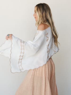A side profile shot of a model in a white embroidered poncho with dramatic bell sleeves layered over a blush midi skirt — soft, romantic, and effortlessly boho.