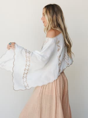 A side profile shot of a model in a white embroidered poncho with dramatic bell sleeves layered over a blush midi skirt — soft, romantic, and effortlessly boho.