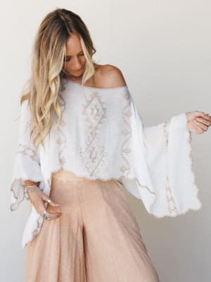 A breezy white embroidered poncho top worn off-shoulder over blush wide-leg pants, styled with silver stacked rings — the kind of outfit that looks equally at home on a cobblestone street or a beach boardwalk.
