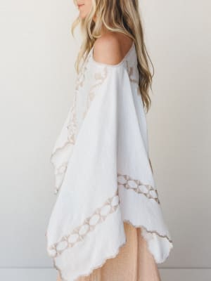 A breezy white poncho with tan embroidered geometric and floral detailing shot in soft side profile — effortless and a little wanderlust, like something you'd throw on over a swimsuit and never take off.