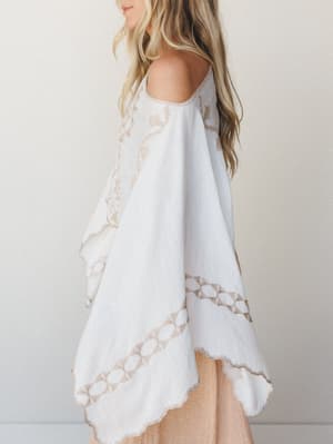 A breezy white poncho with tan embroidered geometric and floral detailing shot in soft side profile — effortless and a little wanderlust, like something you'd throw on over a swimsuit and never take off.