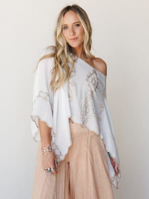 A breezy white poncho with tan embroidery and scalloped trim worn off-shoulder over a blush wide-leg pant — effortless and a little dreamy, like she just landed somewhere warm.