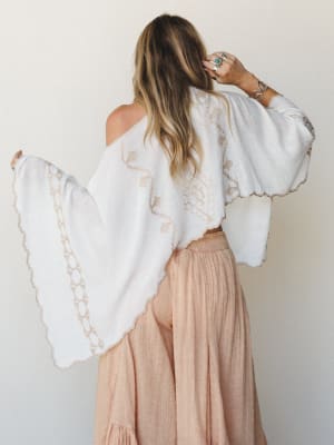 Shot from behind, a model pairs a flowy white embroidered poncho with blush wide-leg pants, silver stacked rings, and a turquoise stone ring — the whole look feels like it was packed for a trip somewhere warm and unhurried.