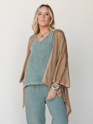A relaxed, earthy layered look — an open-knit camel kimono draped over a dusty blue v-neck top and matching wide-leg pants, styled with stacked stone bracelets and a delicate pendant necklace for that effortlessly collected, boho-everyday feel.
