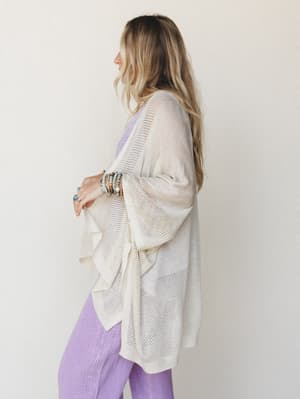 A side-profile shot of a model layering a cream open-knit kimono over a soft lavender set, stacked with beaded bracelets and a turquoise ring — the whole look has that effortless, morning-light energy.
