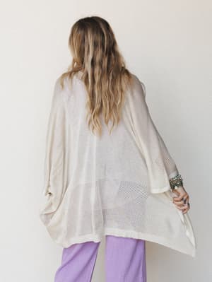 Shot from behind, a model layers an oversized open-knit kimono in warm oatmeal over lavender wide-leg pants, her stacked stone bracelets and rings the only detail you can see — and honestly, it's enough.