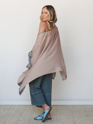 A model shot from the back-turned-to-camera angle, wearing a flowing clay-toned kimono with intricate embroidered border trim peeking out at the sleeves and hem, paired with slate wide-leg pants and bright blue tassel sandals — relaxed, well-traveled