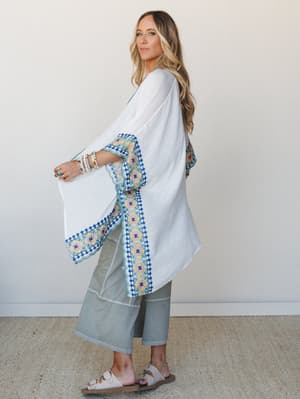 A breezy ivory kimono with vibrant geometric embroidered border trim is styled over sage wide-leg crop pants and flat sandals — the kind of layering piece that makes a simple outfit feel intentional.