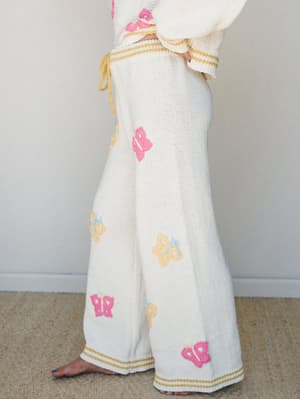 A close-up side shot of wide-leg cream knit pants covered in scattered crochet butterfly appliqués in pink, yellow, and pale blue — finished with a mustard stripe trim at the waist and hem and a drawstring tie.
