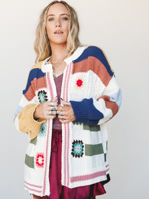 An oversized knit cardigan in cream with colorblock stripes and granny square crochet patches styled open over a burgundy satin skirt — the kind of piece that looks like it has a whole personality before you even put it on.