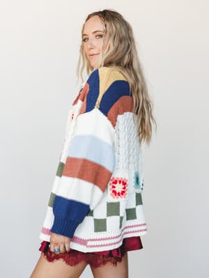 A side-profile shot of a model in an oversized patchwork knit cardigan layered over a burgundy lace-trimmed mini — the kind of sweater that looks like it was made from every good thing at once.