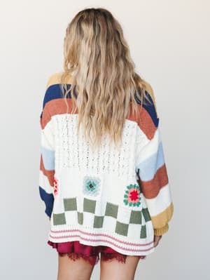 Shot from the back, this oversized cream cardigan layers color-block stripes, granny square crochet florals, and a checkerboard hem into something that feels like your most sentimental thrift-store find — worn over a burgundy lace-trimmed mini for a