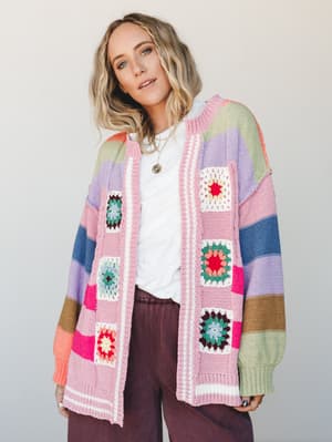 A colorful oversized knit cardigan covered in granny squares and multicolor stripes, worn open over a white tee and dark mauve wide-leg pants — the kind of piece that looks like your most fun thrift store find but make it intentional.