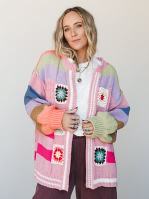 A multicolor striped knit cardigan covered in granny squares is layered over a white tee and worn with burgundy wide-leg pants — the kind of piece that makes a whole room feel warmer just by walking in.