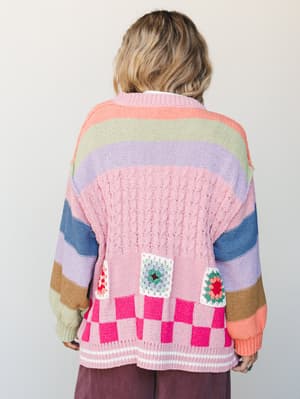 A back-facing shot of an oversized multicolor knit cardigan layered with rainbow stripes, granny square patches, and a checkerboard hem — the kind of sweater that makes a whole personality statement without saying a word.