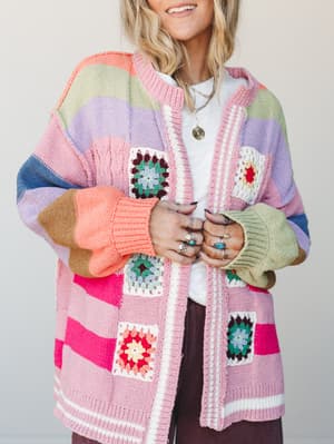 A multicolor striped knit cardigan covered in granny square crochet patches worn open over a white tee — the kind of piece that makes people stop and ask where you got it.