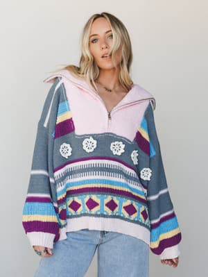 An oversized multicolor knit pullover with a cozy pink fleece-lined collar and half-zip, worn relaxed over light wash jeans — the kind of sweater that makes you want to cancel plans and stay somewhere pretty.