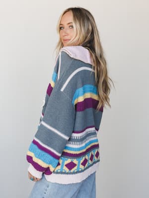A model glances back over her shoulder in a cozy, color-blocked knit pullover with a mix of stripe and geometric Aztec patterns — the kind of sweater that makes a Tuesday feel more interesting.