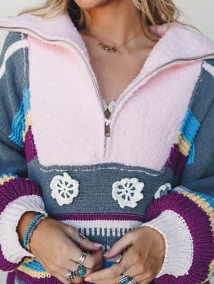 A close-up of a cozy, color-blocked knit pullover with a fluffy pink half-zip collar and crochet flower appliqués, styled with layered silver rings, turquoise bracelets, and a gold 'mama' necklace — the kind of piece that looks like it came from some