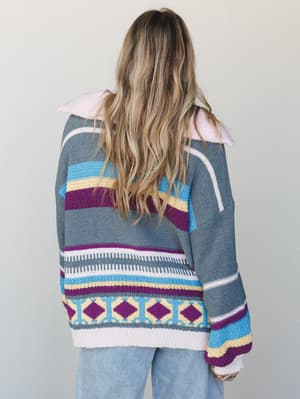 Shot from the back, this oversized multicolor knit pullover layers bold stripe and diamond patterns in grey, purple, and sky blue — worn with light-wash wide-leg jeans for an effortlessly relaxed, cozy-cool vibe.