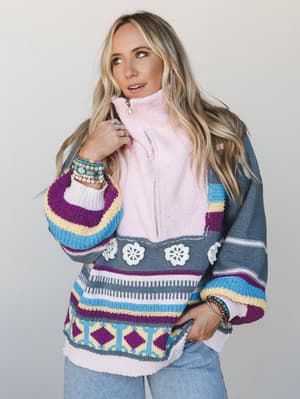 A colorful, oversized knit pullover worn over a blush pink zip-up fleece, layered with stacked beaded bracelets and light wash jeans — the kind of outfit that looks effortless but took exactly the right amount of thought.