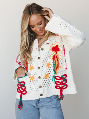 A cream open-knit cardigan covered in bold yarn embroidery and tassel details — floral motifs in red, marigold, and teal give it a handmade, artisan feel that pairs naturally with lived-in light wash denim.