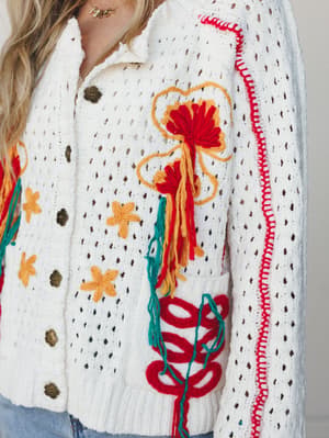 A close-up of a cream open-knit crochet cardigan covered in vibrant yarn embroidery — bold floral motifs, tassels, and looped appliqué in red, golden yellow, and teal spill across the front, finished with antique brass buttons and a red whipstitch tr