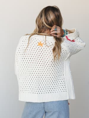 Shot from behind, a model in a cream open-knit cardigan pulls her hair up to show off a small embroidered floral detail on the back — styled with light wash jeans and a stack of turquoise and silver bracelets that feel like they were collected, not c
