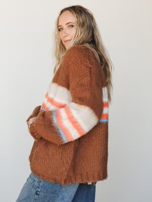 A chunky, fuzzy cardigan in warm brick brown gets a retro lift from color-blocked striped sleeves in coral, cream, and sky blue — styled with relaxed denim for that effortless, lived-in feel.