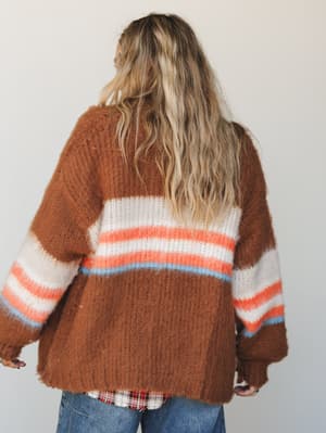 A cozy, oversized striped cardigan in warm brick brown with cream, coral, and sky blue stripe detail — shot from the back, layered over a flannel and jeans for that effortlessly lived-in feel.