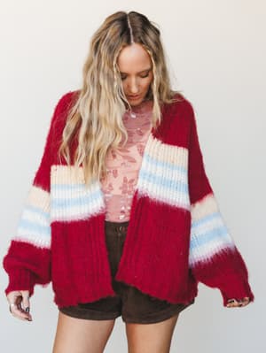 An oversized chunky knit cardigan in cranberry red with cream and sky blue color-block stripes, layered over a sheer floral pink top and dark brown mini shorts — the kind of cozy-cool layering combo that makes fall feel like a personality.