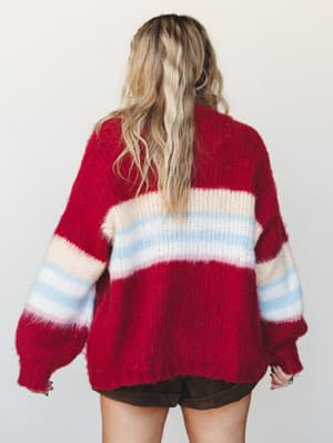 Shot from the back, a chunky striped cardigan in cranberry red with cream, sky blue, and blush stripe detailing gives off that cozy, nostalgic energy — paired with dark brown mini shorts and worn with easy, lived-in confidence.