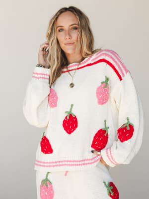 A cream knit sweater covered in chunky embroidered strawberries — some red, some pink — with striped trim at the neckline, cuffs, and hem, styled as a matching set and layered with gold necklaces for a playful, cozy look.
