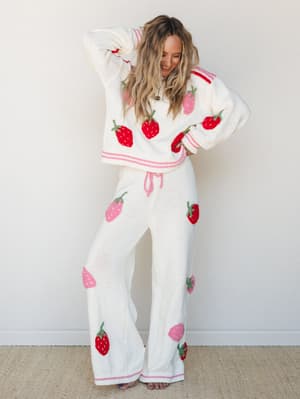 A cream knit matching set covered in embroidered red and pink strawberries — the kind of outfit that makes you smile before you've even put it on, worn here with total ease and a laugh to match.