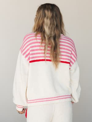 A cream knit sweater shot from the back, with pink stripe detailing across the yoke and hem and a bold red stripe accent — cozy and playful, like something you'd reach for on a slow Saturday morning.