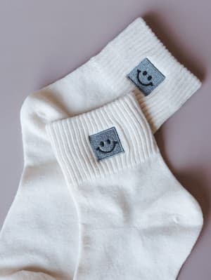 A close-up of two ivory crew socks layered casually against a muted lavender background, each cuff featuring a small embroidered smiley face patch — the kind of little detail that makes getting dressed feel like a good idea.