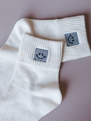Two ivory crew socks laid casually on a muted lavender surface, each with a small embroidered smiley face patch on the ribbed cuff — the kind of detail you notice and immediately want.