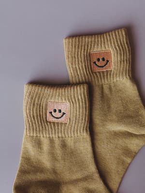 A pair of mustard yellow crew socks laid flat against a soft lavender background, each cuff detailed with an embroidered smiley face patch — the kind of small, happy thing that makes the whole outfit.