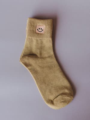 A single mustard yellow crew sock laid flat against a soft lavender-grey background, with a small embroidered smiley face patch on the folded cuff — the kind of little-detail accessory that makes an outfit feel intentional.