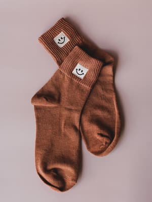 A pair of rust-toned ribbed crew socks laid casually on a blush surface, each cuffed ankle sporting a small white embroidered smiley face patch — the kind of detail that makes an ordinary Tuesday feel a little more intentional.