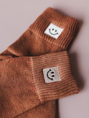 A close-up of two pairs of rust-toned marled crew socks with embroidered smiley face patches on the cuffs — one happy, one upside-down — layered casually against a soft blush background.