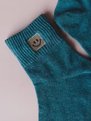 A close-up of heathered teal crew socks with a ribbed cuff and a small embroidered smiley face patch — the kind of tiny detail that makes getting dressed feel a little more fun.