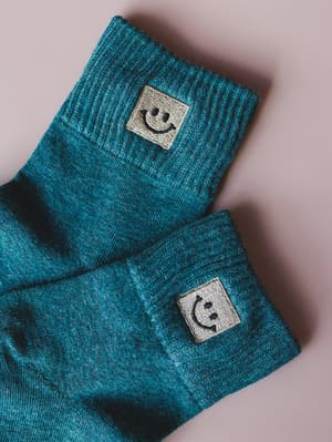 A close-up of two teal marled crew socks laid casually on a blush surface, each cuffed to show off a small embroidered smiley face patch — the kind of detail that makes getting dressed feel a little more fun.