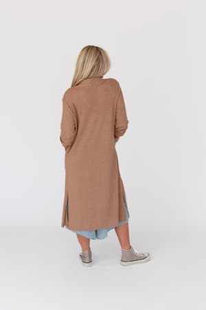 A woman with long blonde hair faces away, wearing The Nest Jayde Waffle Knit Cardigan in taupe over a blue dress with matching taupe sneakers, set against a plain white background.