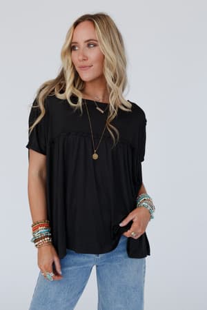 A woman with long blonde hair wears the Sunday Morning Babydoll Top - Black, styled with blue jeans, layered necklaces, and colorful bracelets, standing against a plain light background and looking slightly to the side.