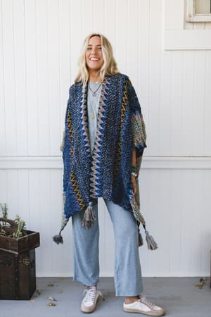 A woman with long blonde hair smiles while wearing the colorful Desert Dreams Crochet Ruana - Cobalt with tassels, paired with light blue wide-leg pants and white sneakers, standing by a white wall and plants for a boho vibe.