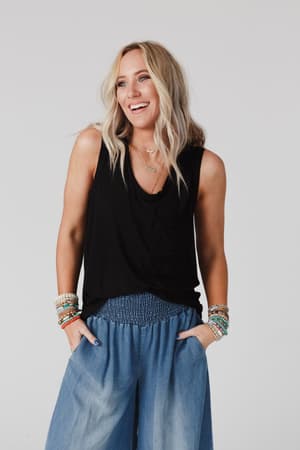 A smiling woman with wavy blonde hair wears The Nest Shelli Scoop Neck Tank Top in black and wide-leg blue pants, accessorized with colorful bracelets, hands in her pockets, standing against a plain light background.