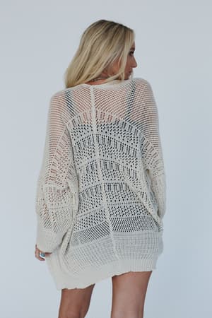A woman with long blonde hair stands facing away from the camera, wearing the Brenden Knit Netted Cardigan in Ivory over dark undergarments, against a plain white background.