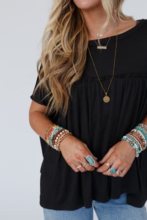 A woman with wavy blonde hair wears the Sunday Morning Babydoll Top - Black, paired with blue jeans, layered necklaces, beaded bracelets, and turquoise rings. Her face is partially out of frame.